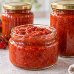 Лютеница от печени чушки и домати за килера Три буркана с домашна лютеница от печени червени чушки и домати, подредени до печени чушки и пресен домат.