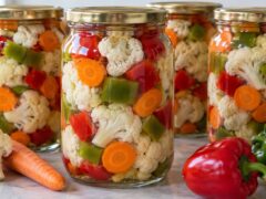 Четири буркана с маринован карфиол, моркови и червени и зелени чушки, подредени до пресни зеленчуци.