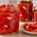 Маринована червена капия за зимата Три буркана с маринована печена червена капия и чесън, с порция чушки и скилидки чесън в чиния.